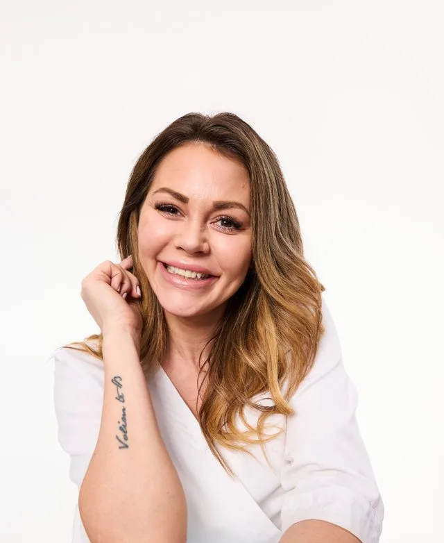 Camilla, hudterapeut, med langt, bølget blondt hår og naturlig sminke, iført en hvit frakk med en Skin City-logo på venstre side, mot en hvit bakgrunn.