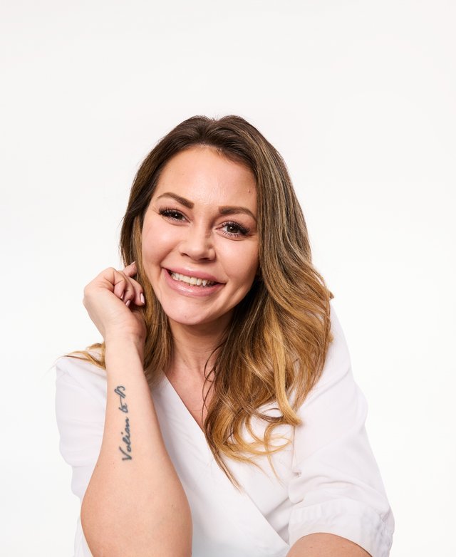 Camilla, ihoterapeutti, jolla on pitkät, lainehtivat vaaleat hiukset ja luonnollinen meikki, pukeutuneena valkoiseen takkiin, jossa on Skin City -logo vasemmalla puolella, valkoista taustaa vasten.
