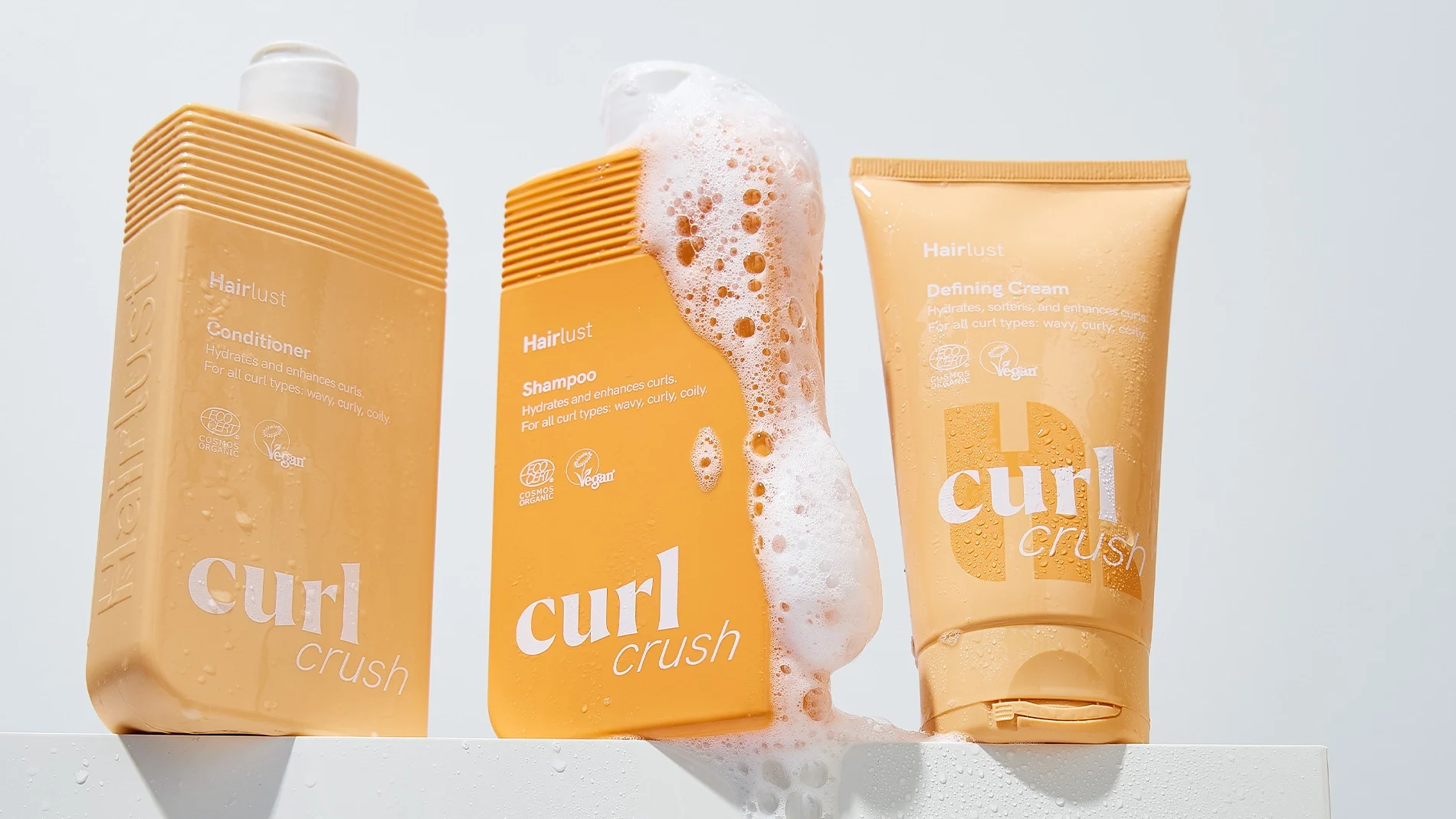 Tre Hairlust Curl Crush-produkter för lockigt hår står på en vit yta: Conditioning Wash, Shampoo och Defining Crème. Förpackningarna är i beige och orange toner, och vitt skum rinner över shampooflaskan. Synlig text inkluderar “Hairlust”, “Conditioner Wash”, “Shampoo”, “Defining Crème”, “Vegan” och “curl crush”.