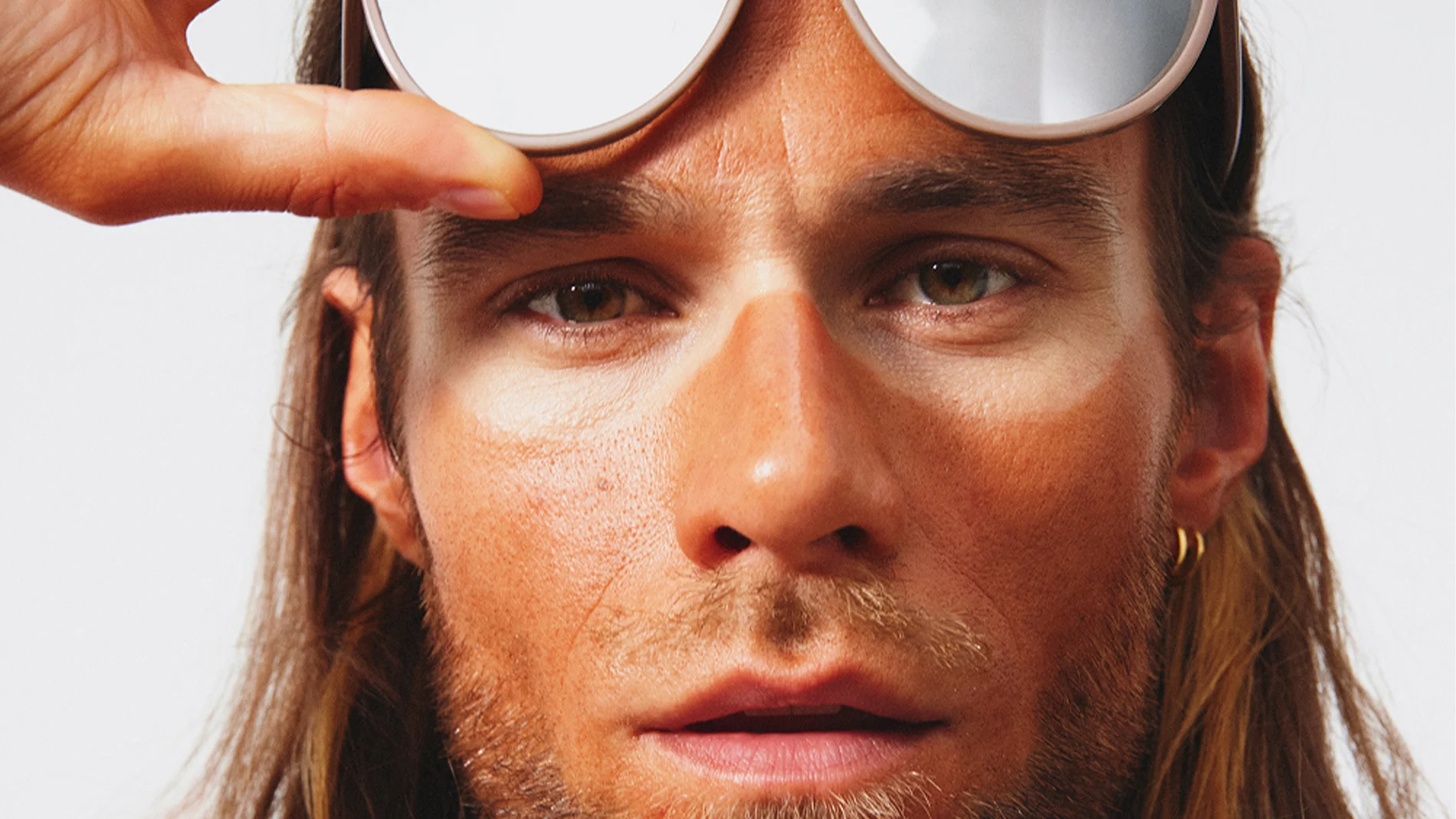 En person håller upp ett par stora, spegelblanka solglasögon ovanför ögonen, vilket avslöjar tydliga linjer efter solskydd och en markerad kontrast mellan solbrända och oskyddade partier i ansiktet, med fokus på hudvård och SPF.