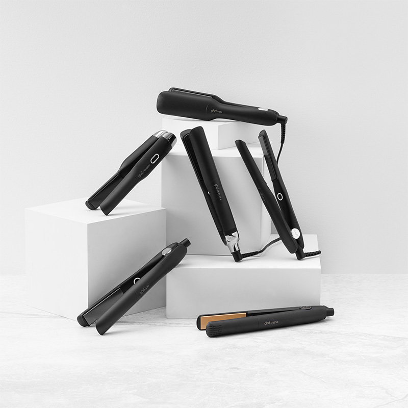 En samling svarte GHD-strykejern er stilfullt arrangert på hvite kuber mot en lysegrå bakgrunn. De flate jernene har elegante detaljer og er plassert i ulike vinkler for å vise design og funksjon.