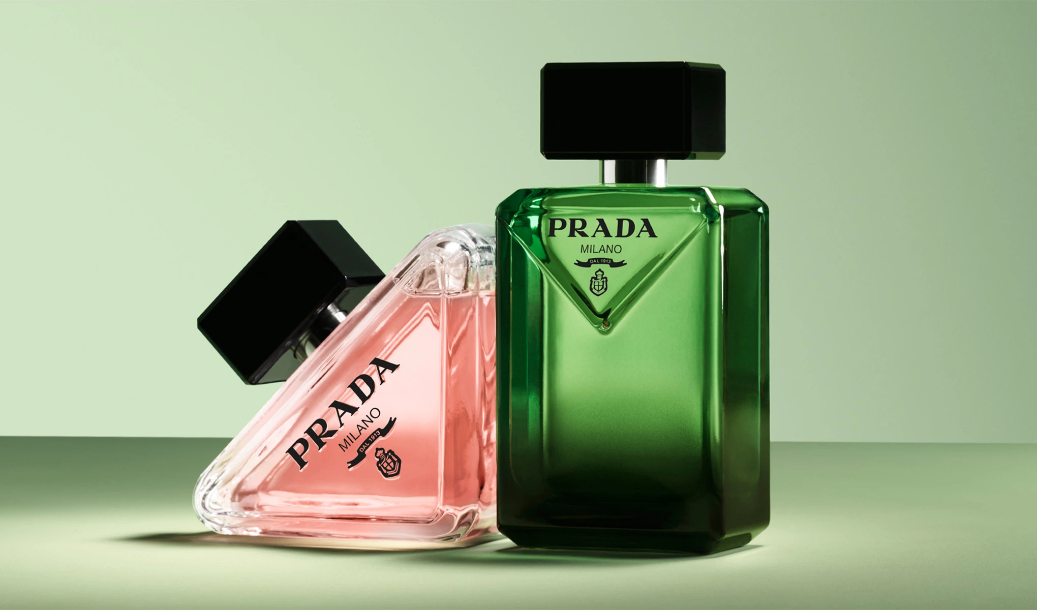 Kaksi Prada Milano -hajuvesipulloa vaaleanvihreällä taustalla. Vasemmalla vaaleanpunainen pullo, joka makaa kyljellään, ja oikealla pystyssä oleva vihreä pullo. Molemmissa pulloissa on musta korkki.