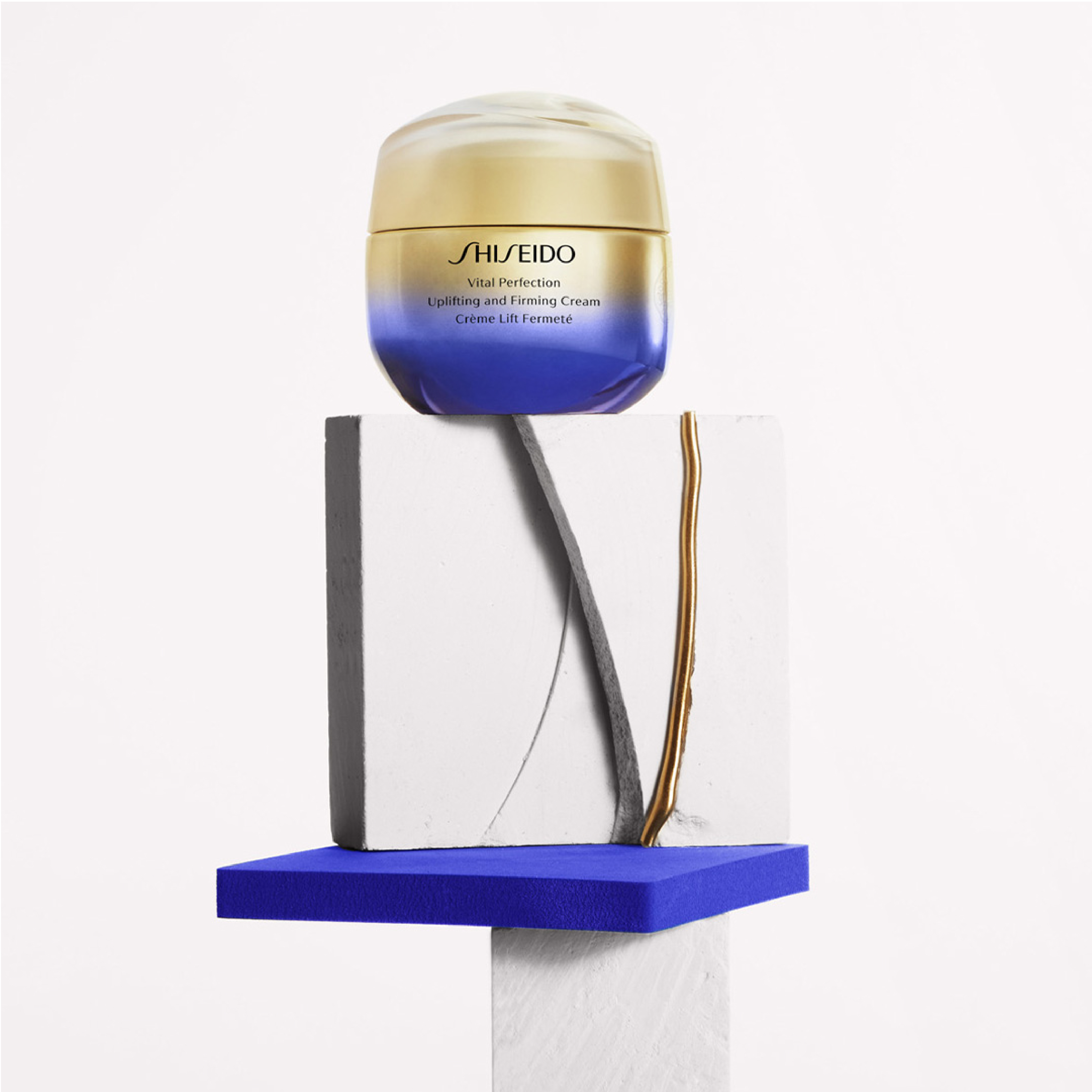 Shiseido-voide kultaisessa ja sinisessä purkissa, asetettuna valkoiselle ja siniselle jalustalle mustan oksan vieressä.
