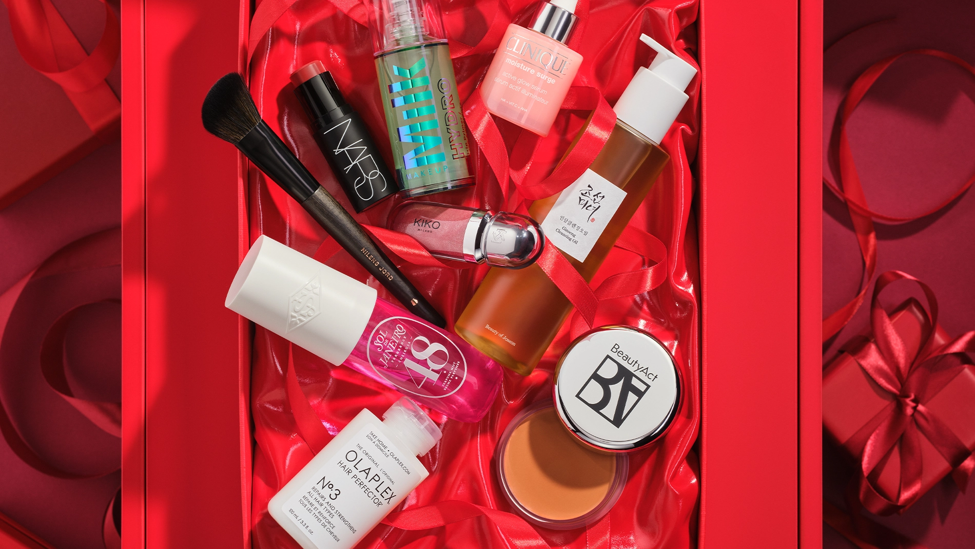 Bild som visar flera skönhetsprodukter arrangerade i en röd presentförpackning. Produkterna inkluderar en svart flaska med NARS foundation, en brun flaska med SKIN VEIL, en rosa flaska med Clinique Moisture Surge, en vit flaska med pump, en burk med BeautyAct bronzer, en flaska med Olaplex Hair Perfector No.3, en makeupborste, och flera andra skönhetsprodukter. Bakgrunden är röd.