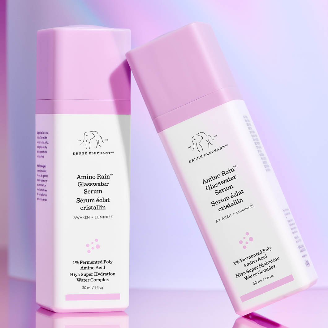 Kaksi vaaleanpunaista ja valkoista Drunk Elephant Amino Rain Glasswater Serum -pulloa seisoo vaalealla, pastellisävyisellä taustalla. Teksti "AWAKEN + LUMINIZE" ja tuotteen yksityiskohdat näkyvät pulloissa.