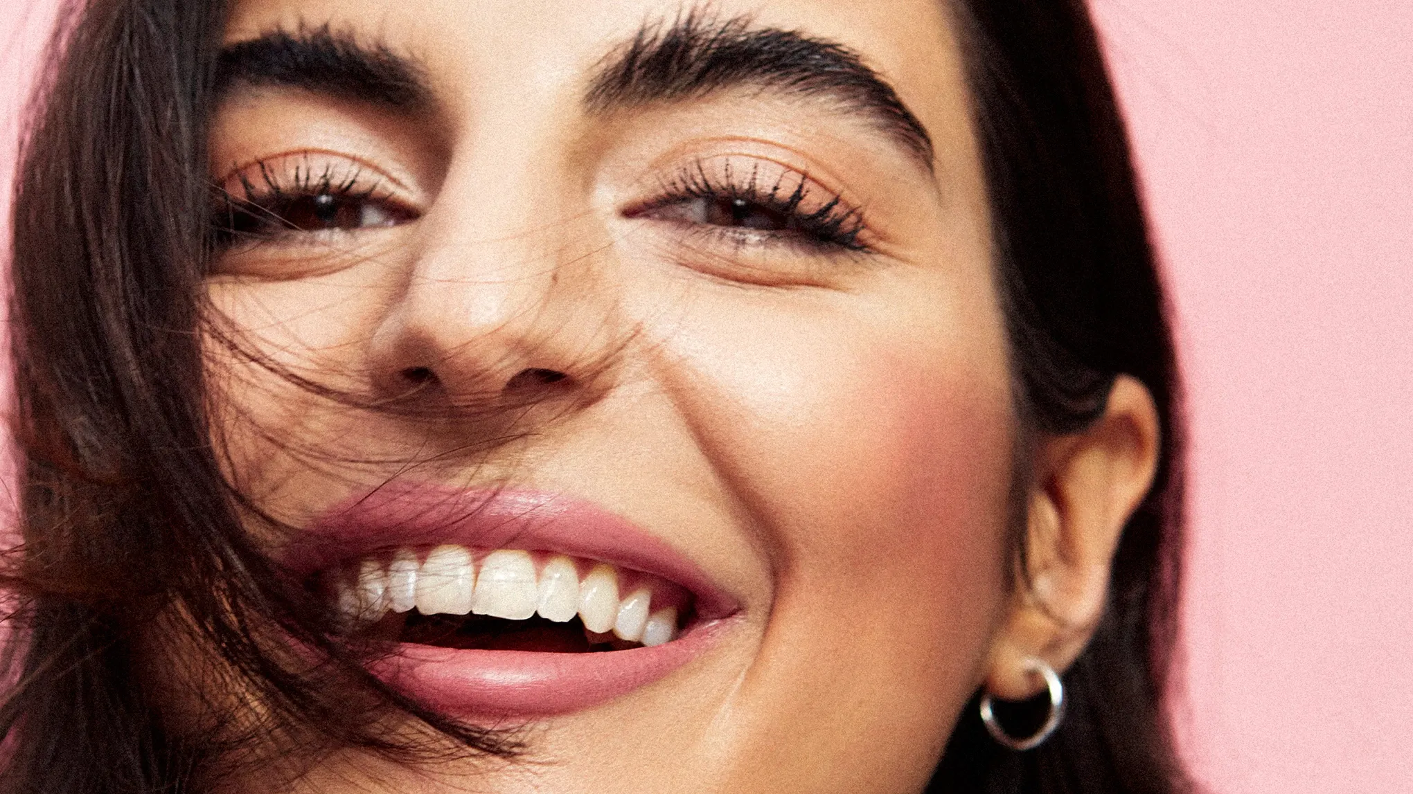 Närbild på leende kvinna med glansig hud, rosiga kinder och naturlig makeup mot rosa bakgrund.