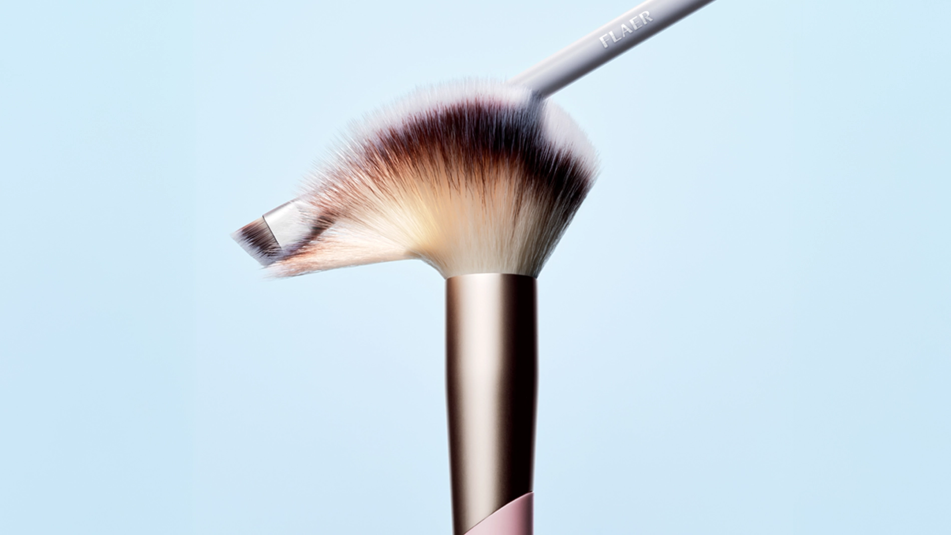 Två sminkborstar mot ljusblå bakgrund, en stor fluffig borste med beige och mörka strån samt metallisk kopparfärgad hylsa, och en mindre snedställd borste placerad ovanpå. Närbild som visar borsthuvudens form och struktur.