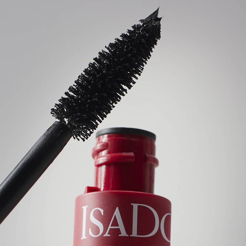 En öppen mascara från IsaDora. Mascaraborsten är täckt med svart mascara och hålls ovanför den röda förpackningen med texten "IsaDora". Bakgrunden är ljusgrå.