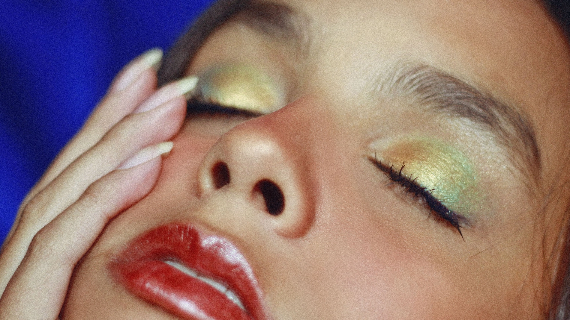 Närbild av ett ansikte med stängda ögon, glänsande röd läppglans och metallisk ögonskugga i gröna och gyllene nyanser.
