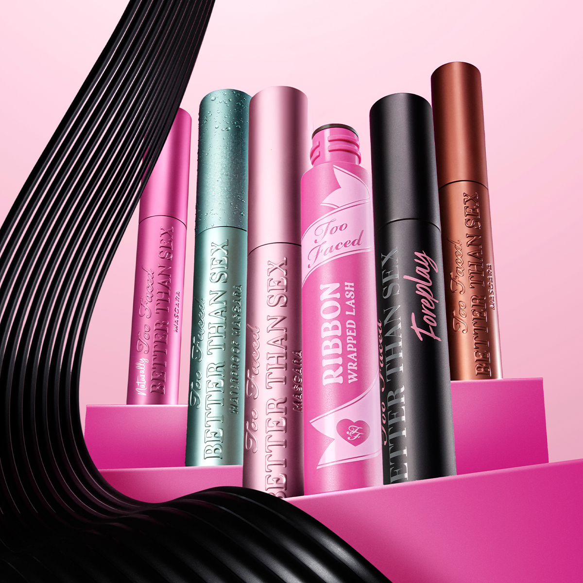 Seks fargerike Too Faced-maskaraer mot en rosa bakgrunn. Fra venstre til høyre: rosa, grønn, lys rosa, rosa med båndmønster, svart og bronsefarget.