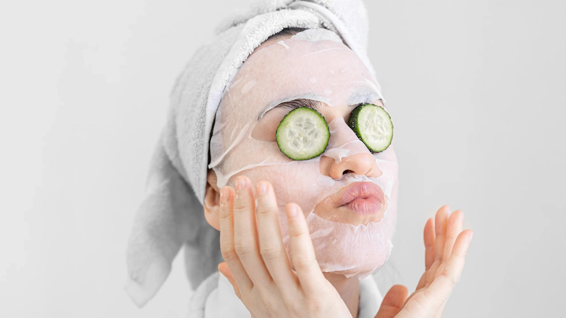 Kvinna med håret i vit handduk samt ansiktsmask och gurkor på ögonen