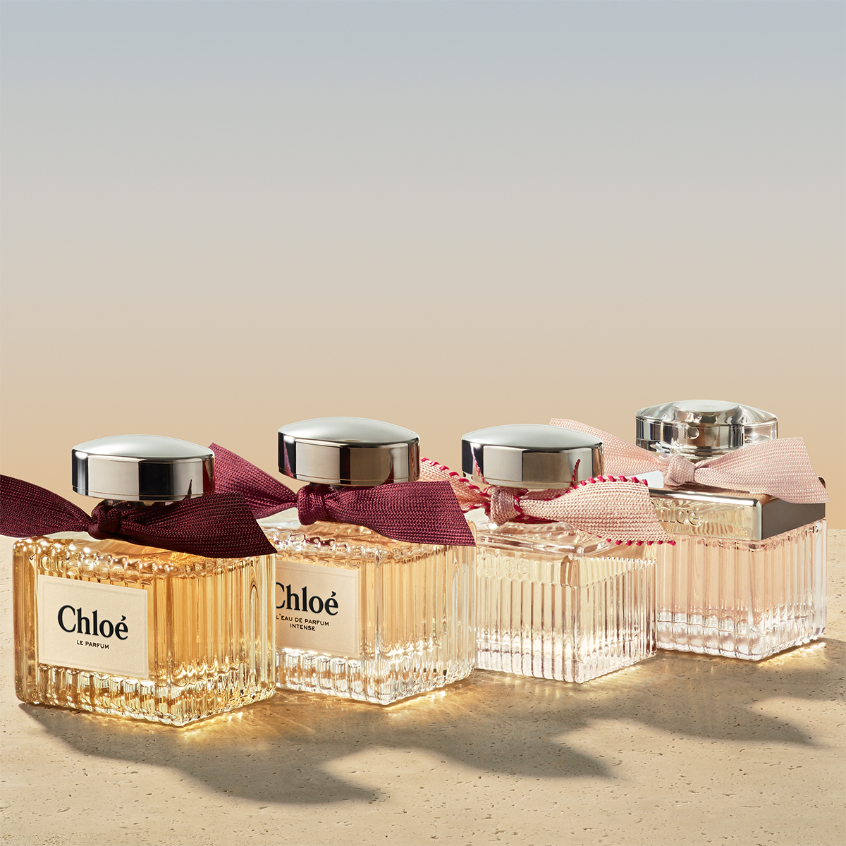Tre parfymeflasker fra Chloé. Den ene flasken er dekorert med en mørkerød sløyfe og har en sølvfarget kork. De to andre er dekorert med en lys rosa sløyfe. De er plassert på en lys flate med myk belysning som skaper refleksjoner. Bakgrunnen er lys og nøytral.