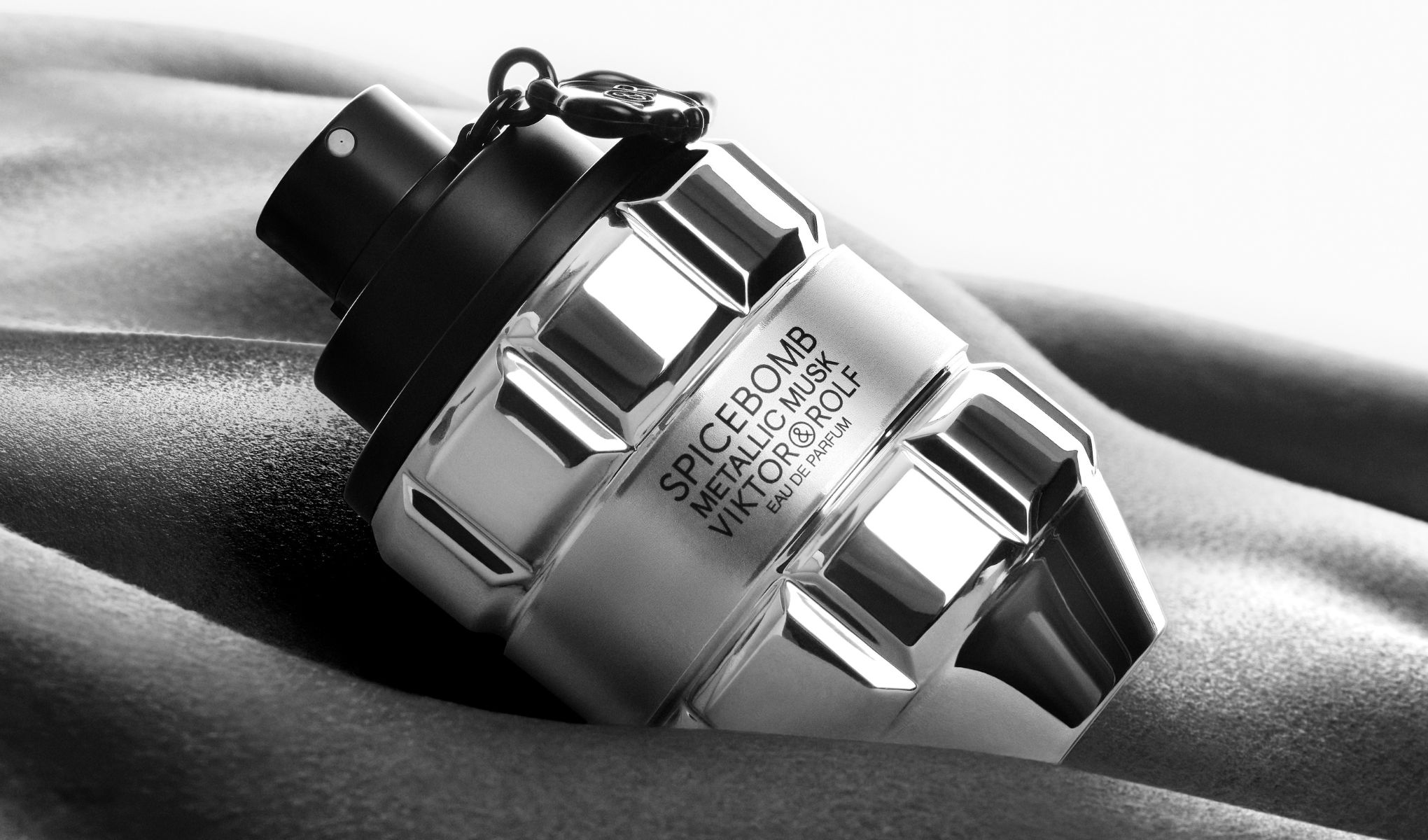 Viktor & Rolf Spicebomb Metallic Musk-parfyme er avbildet i en metallisk flaske som ligner en håndgranat. Flasken hviler på en myk, bølgende overflate.