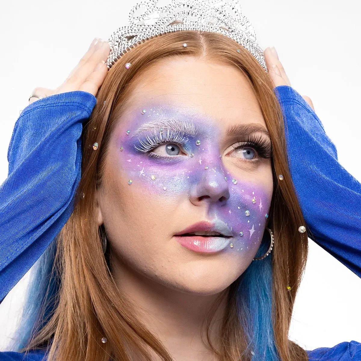 En person med galaxinspirerad makeup i lila, blå och silver med vita fransar, glittrande stenar och målade stjärndetaljer håller en glittrande tiara på huvudet, iförd en blå topp.