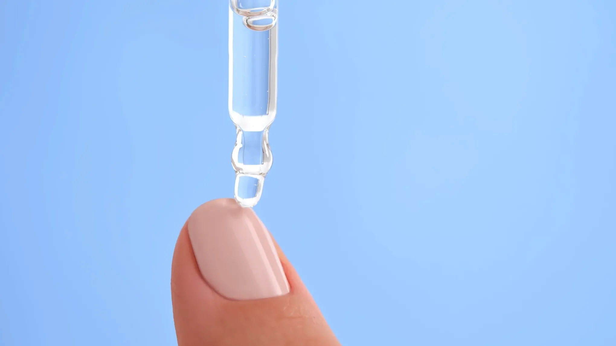 Nærbilde av en dråpe gjennomsiktig serum som påføres en velmanikyrert fingertupp mot en lys blå bakgrunn. Serumet dryppes fra en pipette, og fingertuppen har en lys rosa neglelakk.