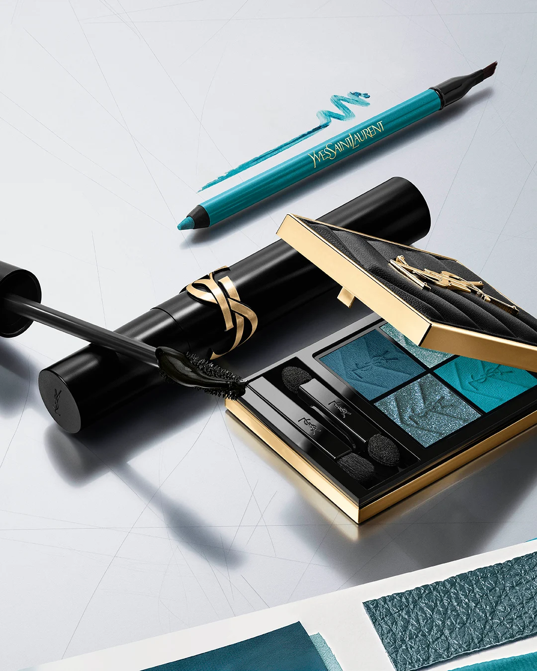 En person med turkis øyesminke og skimrende highlighter holder en åpen Yves Saint Laurent-øyenskyggepalett med fire blå og turkise nyanser foran ansiktet. Personen bærer gullfargede øredobber, og paletten inneholder også applikatorer.