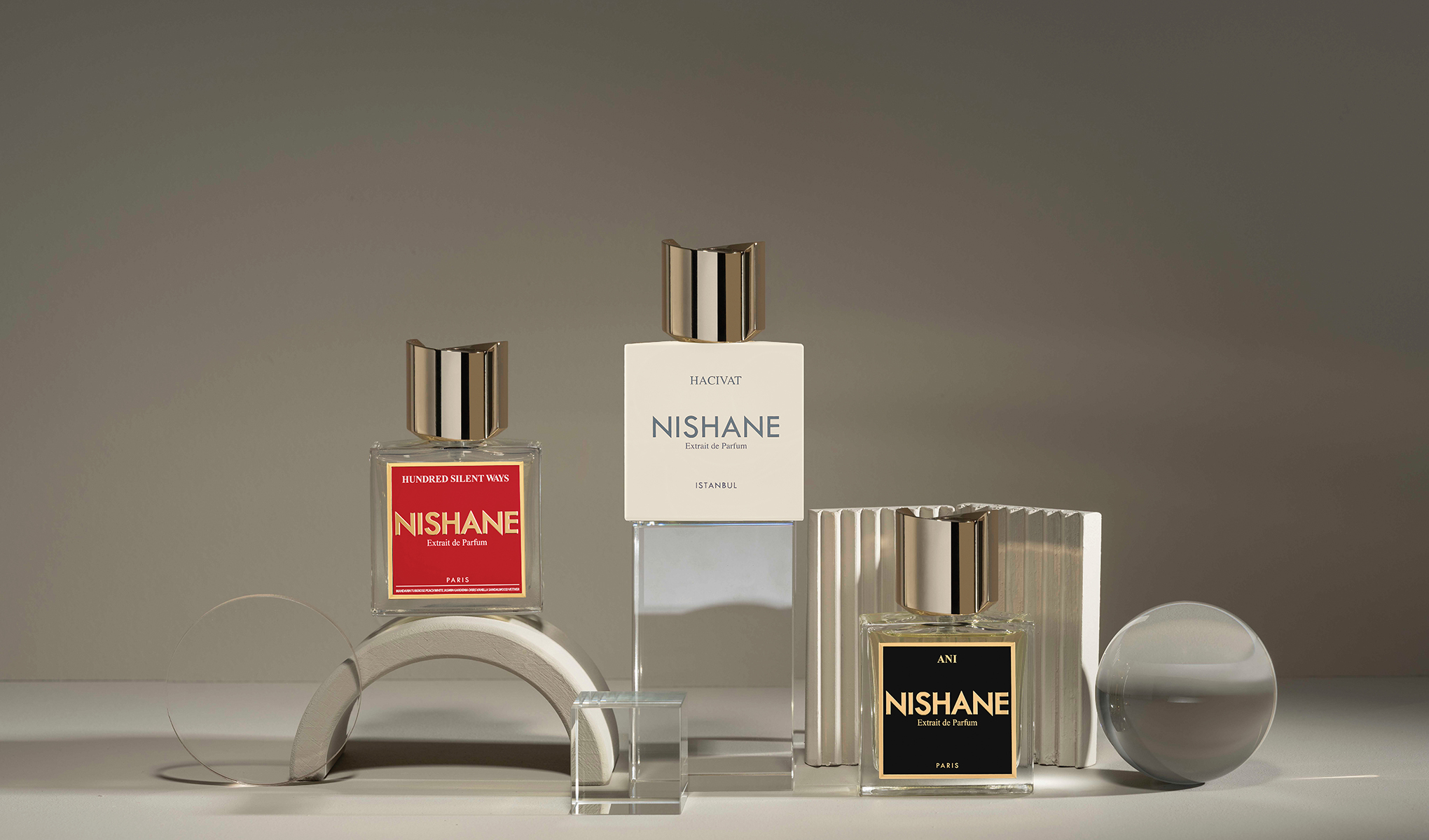 Tre Nishane-parfymer står utstilt på et bord. Den til venstre med rød etikett, den i midten med hvit etikett og den til høyre med svart etikett. Flaskene har elegante, gylne korker og er omgitt av geometriske dekorasjoner.