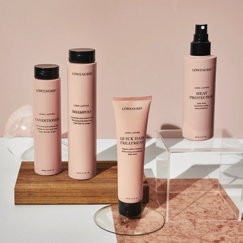 Fyra hårvårdsprodukter från Löwengrip står uppradade mot en rosa bakgrund. Alla produkter har rosa förpackningar med svart text och svarta lock. Produkterna är placerade på en träplatta och en genomskinlig kub.