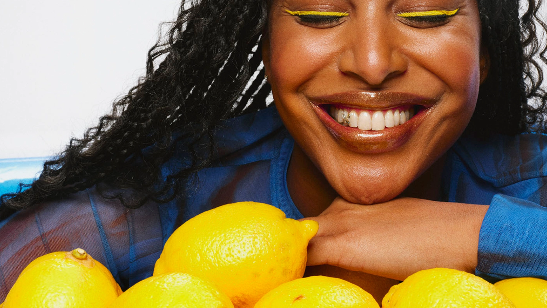 Person med gul eyeliner, blank lipgloss og blå topp lener seg over flere gule sitroner i et fargerikt beautybilde med fokus på makeup som eyeliner og lipgloss.