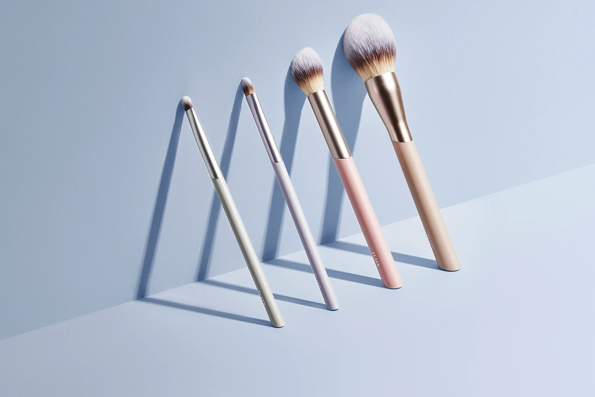 Neljä FLAER-merkin meikkisivellintä on rivissä vaaleansinistä taustaa vasten. Siveltimissä on eri kokoja ja värejä, kuten valkoinen, vaalean violetti, vaaleanpunainen ja beige, ja niissä on hopeisia ja kultaisia yksityiskohtia. Siveltimien varjot luovat tyylikkään ja esteettisen vaikutelman.