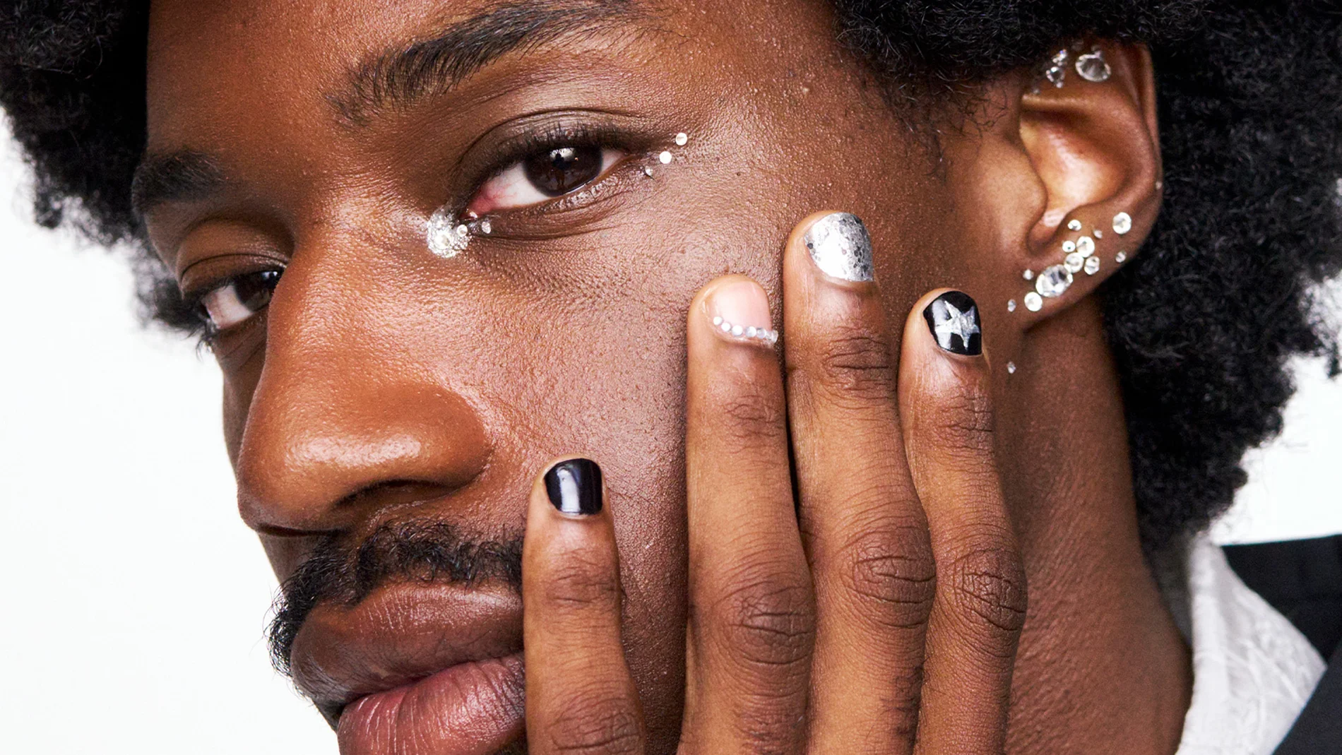Et nært portrett viser en person med dyp hudtone og naturlig krøllete hår, der ansiktet hviler mot hånden i hovedfokus. Neglene er dekorert med en kombinasjon av svart lakk, sølvglitter og kunstneriske detaljer, inkludert et stjernemotiv og små krystaller som gir et eksklusivt og trendy uttrykk. Rundt øynene sees skimrende sminke og subtile glitresteiner langs kinnbenet, som fremhever et kreativt og festinspirert beauty-look. Bildet oser av moderne stil, individualitet og fokus på detaljert negledesign.