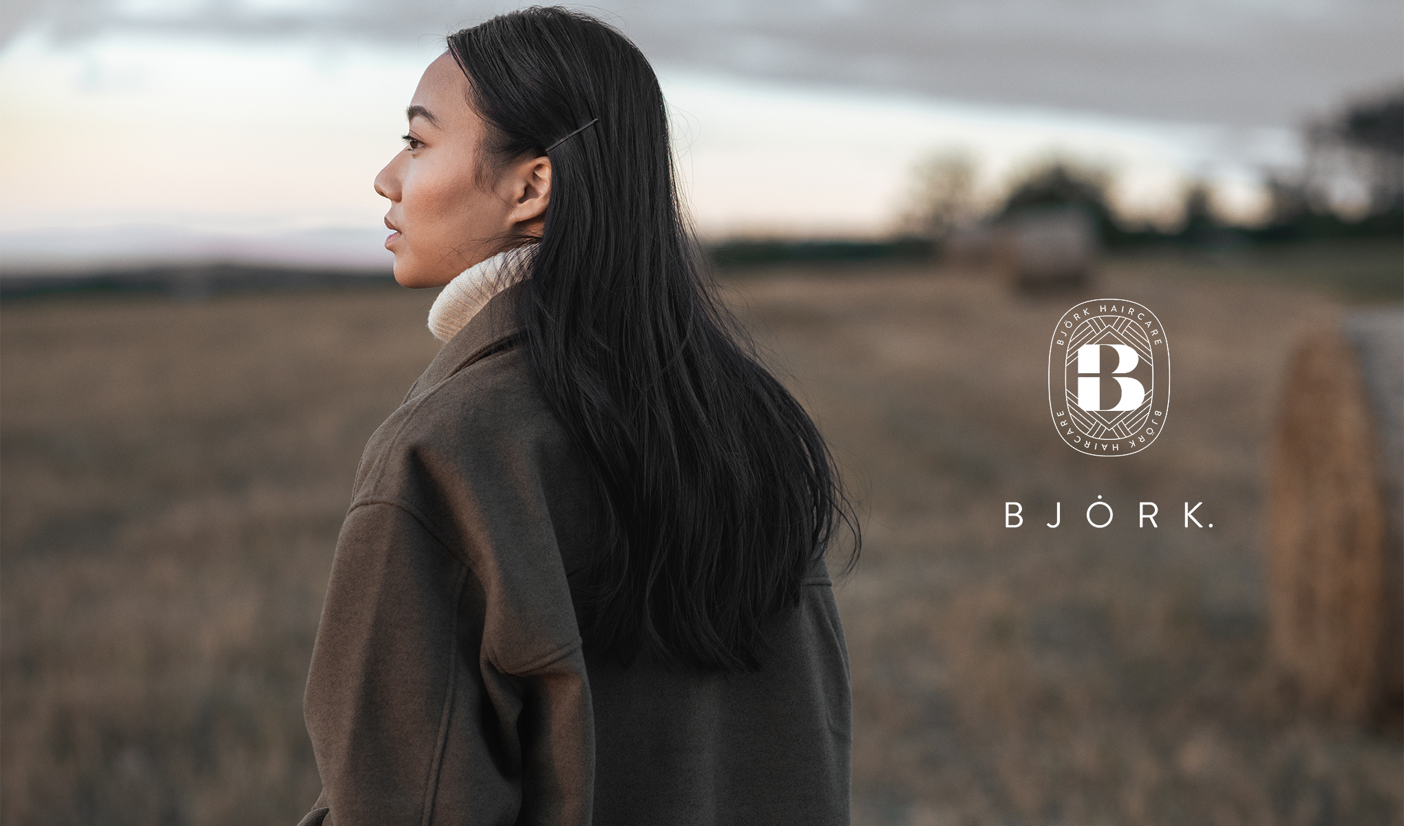 Henkilö seisoo ulkona pellolla syksyisessä ympäristössä. Hänellä on pitkät, tummat hiukset ja hän on pukeutunut ruskeaan takkiin. Kuvassa on myös Björk Haircare -logo ja teksti "BJÖRK."