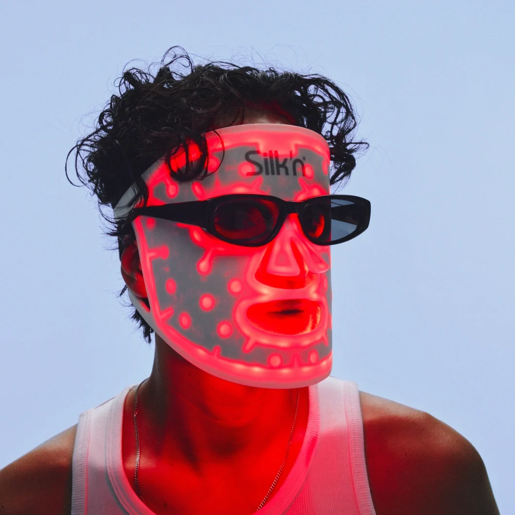 En person med mörkt lockigt hår bär en upplyst Silkn LED-mask och svarta glasögon mot ljusblå bakgrund. Den röda LED-belysningen lyser genom masken och ansiktet.