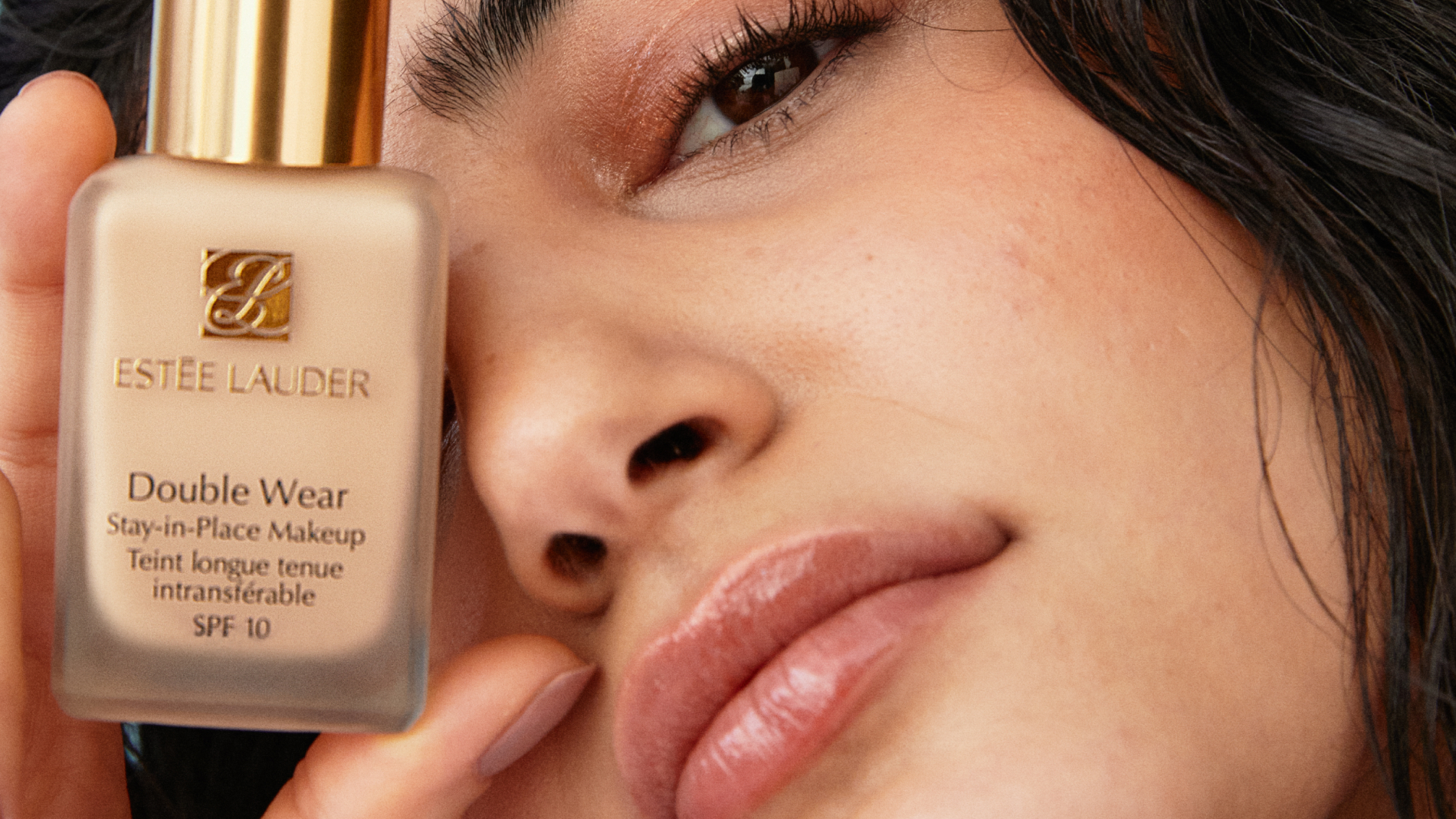 Käsi pitelee Estée Lauder Double Wear Stay-in-Place Makeup SPF 10 -pulloa. Pullossa on kultainen korkki ja pullon teksti on selvästi näkyvissä.