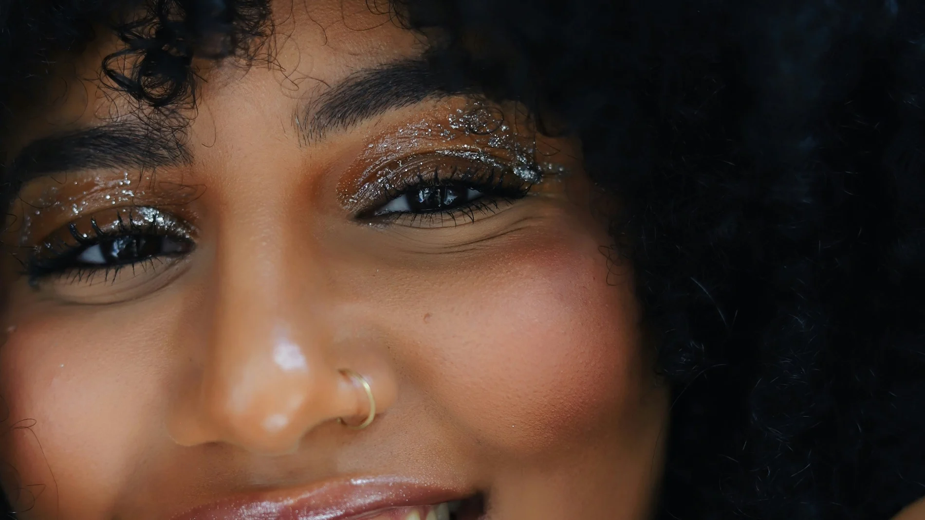 Närbild på en person med mörkt lockigt hår, glittrande ögonmakeup, markerade ögonfransar och glansiga läppar. Personen ler och bär en guldfärgad näsring.