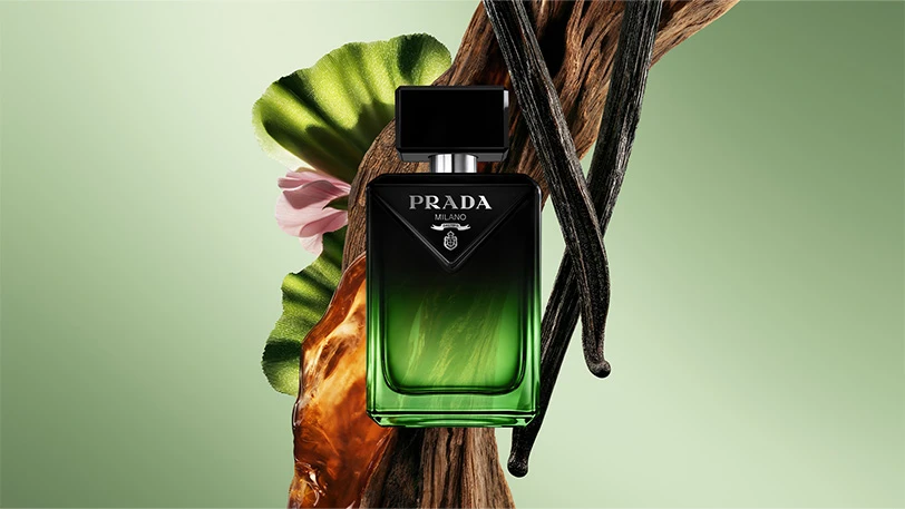 Prada Milano parfymflaska, grön-svart gradient. Omgiven av trä, blad, rosa blomma och vaniljstänger.