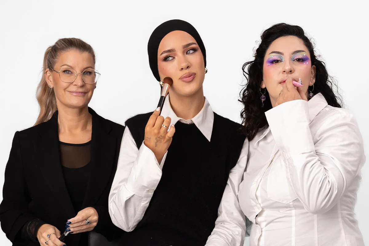 Tre personer sitter tillsammans, där en håller en makeupborste och de andra bär kreativa makeuplooker – en med markerad bas och eyeliner samt en med färgstark ögonmakeup med glitterdetaljer.