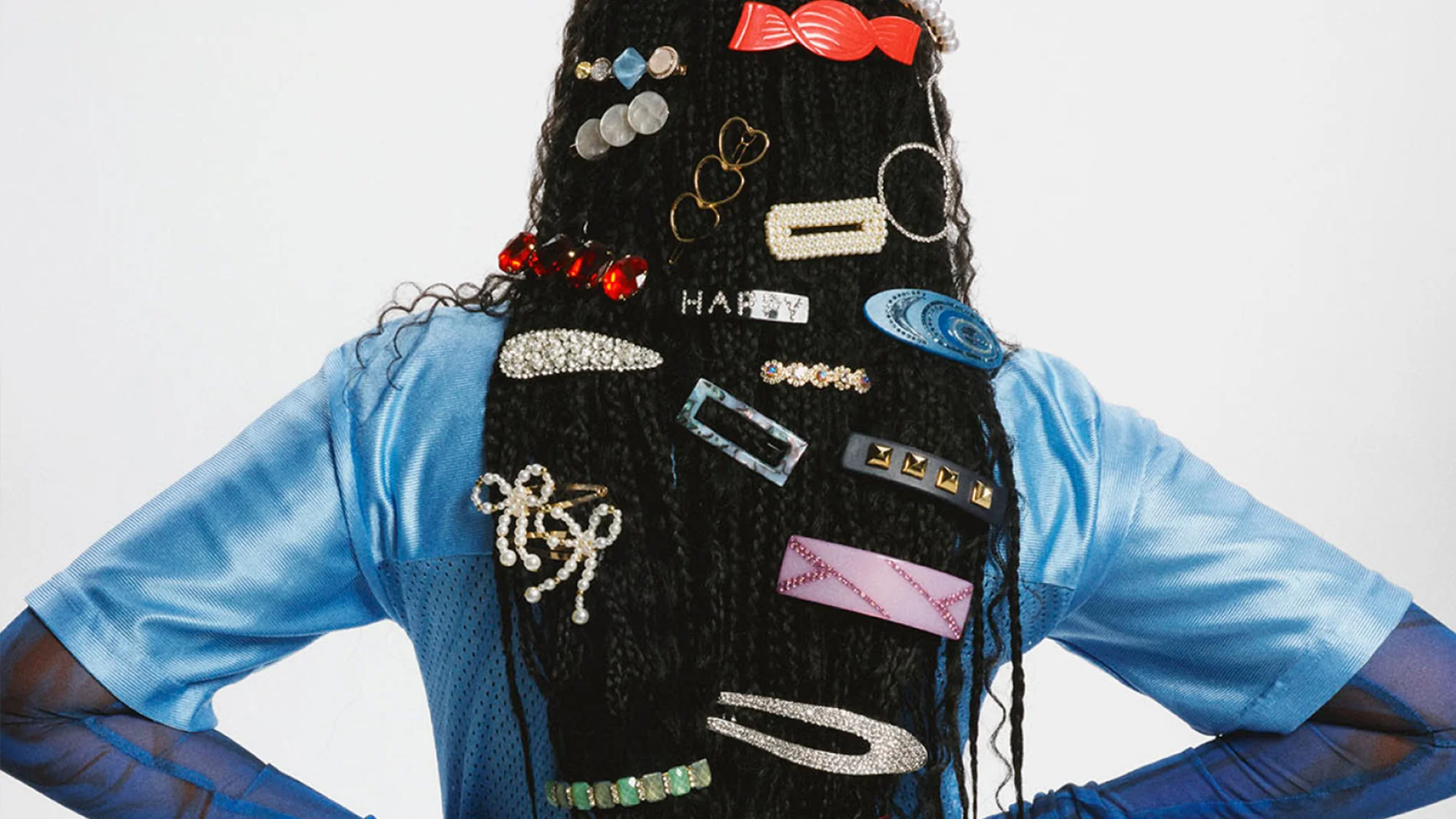 En person sedd bakifrån med långt tvinnat hår fullt dekorerat med färgstarka hårklämmor, pärlspännen, glittrande clips och formade accessoarer i olika stilar, bärandes en blå topp med meshärmar.