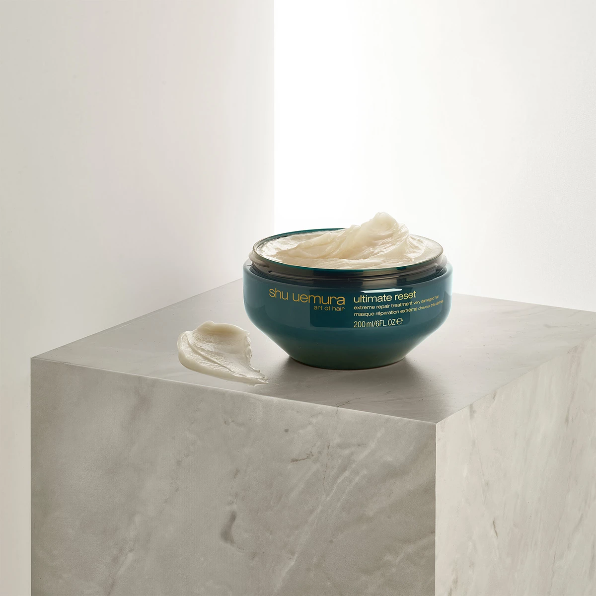 Purkillinen Shu Uemura Ultimate Reset -hiusnaamiota, asetettuna valkoiselle marmorikuutiolle. Osa hiusnaamiosta on levitetty purkin viereen.