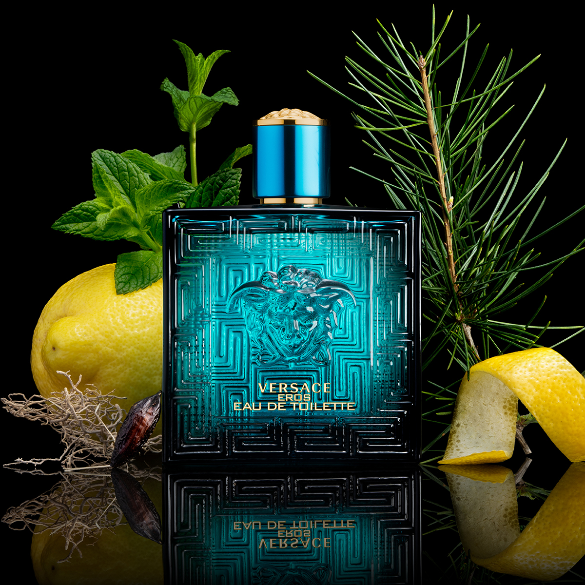 Turkoosi Versacen hajuvesipullo, jossa on kultainen korkki, jota ympäröi sitruuna, sitruunankuori, minttu, männynoksat ja mausteet mustalla pohjalla. Pullossa on teksti ”Versace Eros Eau de Toilette”.