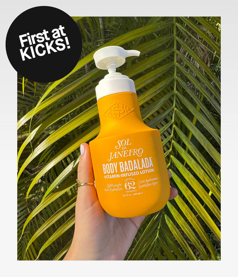 Käsi pitää Sol de Janeiro Body Badalada -pulloa vihreää palmulehteä vasten. Musta tarra, jossa lukee "First at KICKS!"
