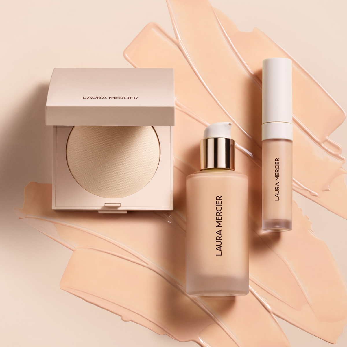 Tre Laura Mercier-produkter: ett kompakt-puder, en flytande foundation och en concealer. Produkterna är placerade på en ljus bakgrund med foundation utsmetad bakom dem.