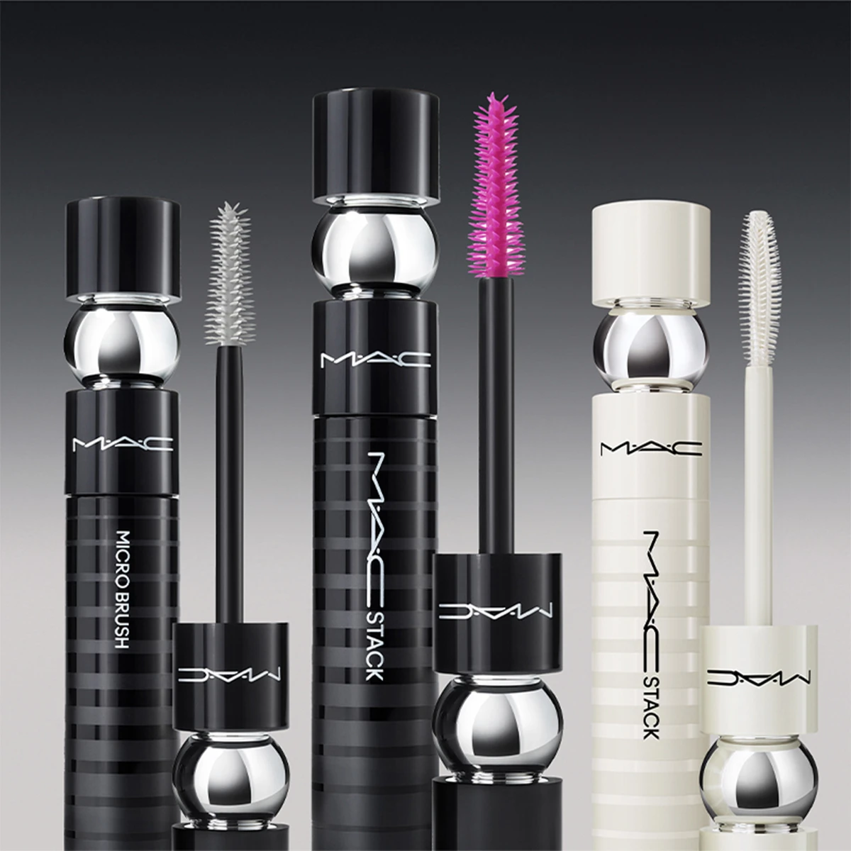 Fire forskjellige mascaraer fra MAC Cosmetics er stilt opp mot en mørkegrå bakgrunn. Emballasjen er svart og hvit med sølvfargede detaljer, og MAC-logoen er godt synlig på hver mascara. Børstene er i forskjellige farger.