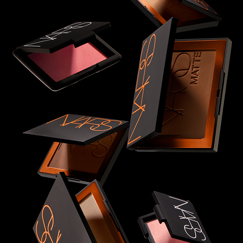 Flera NARS makeup-förpackningar i olika nyanser, svävande mot en svart bakgrund. Kompakterna har svarta höljen med NARS-logotypen tryckt i orange på locket. Inuti syns olika färger av rouge och bronzer, från rosa till bruna toner. 