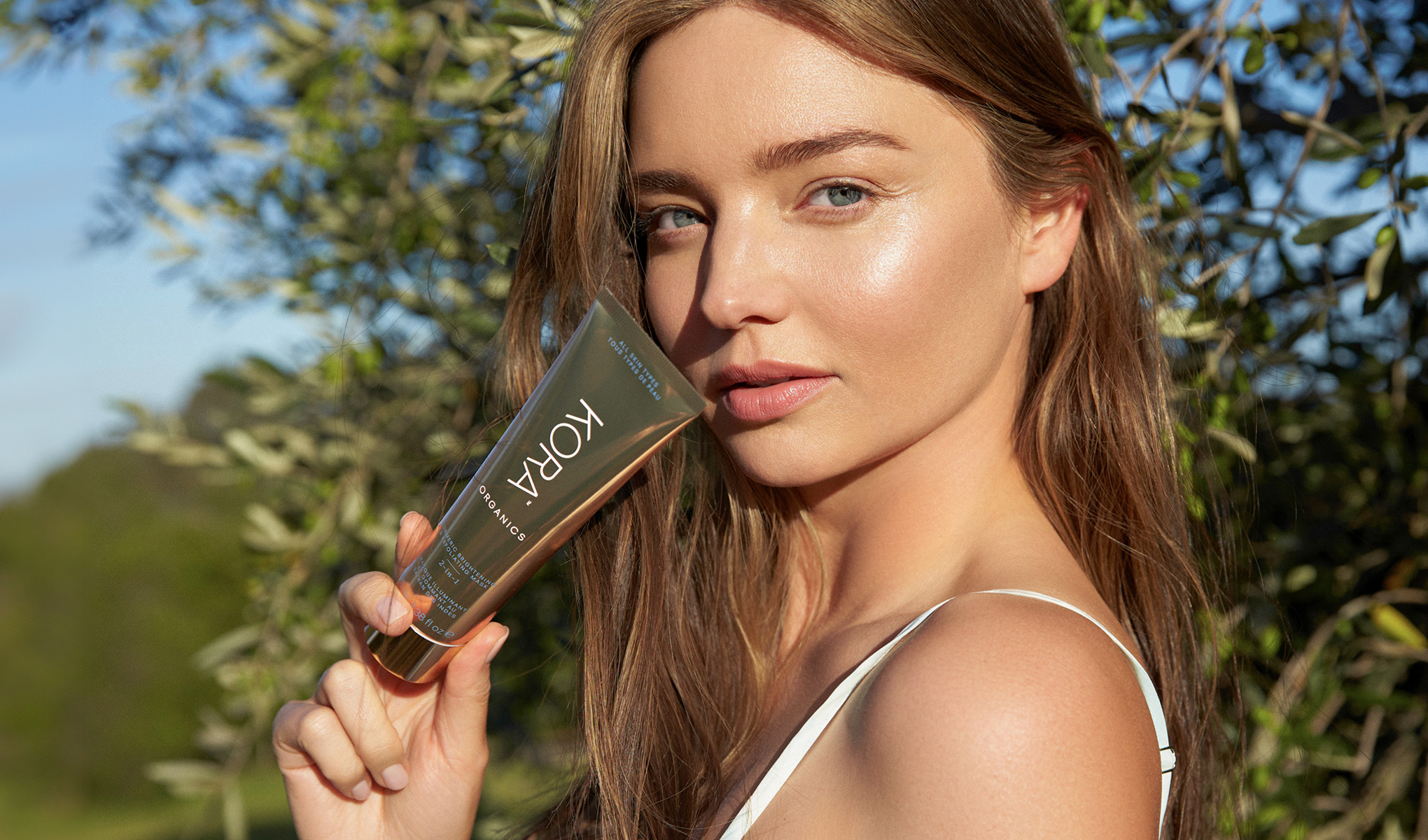 En person håller en tub med KORA Organics produkt. Personen har långt brunt hår och bär en vit topp. Bakgrunden visar grönska och träd under en klar himmel.