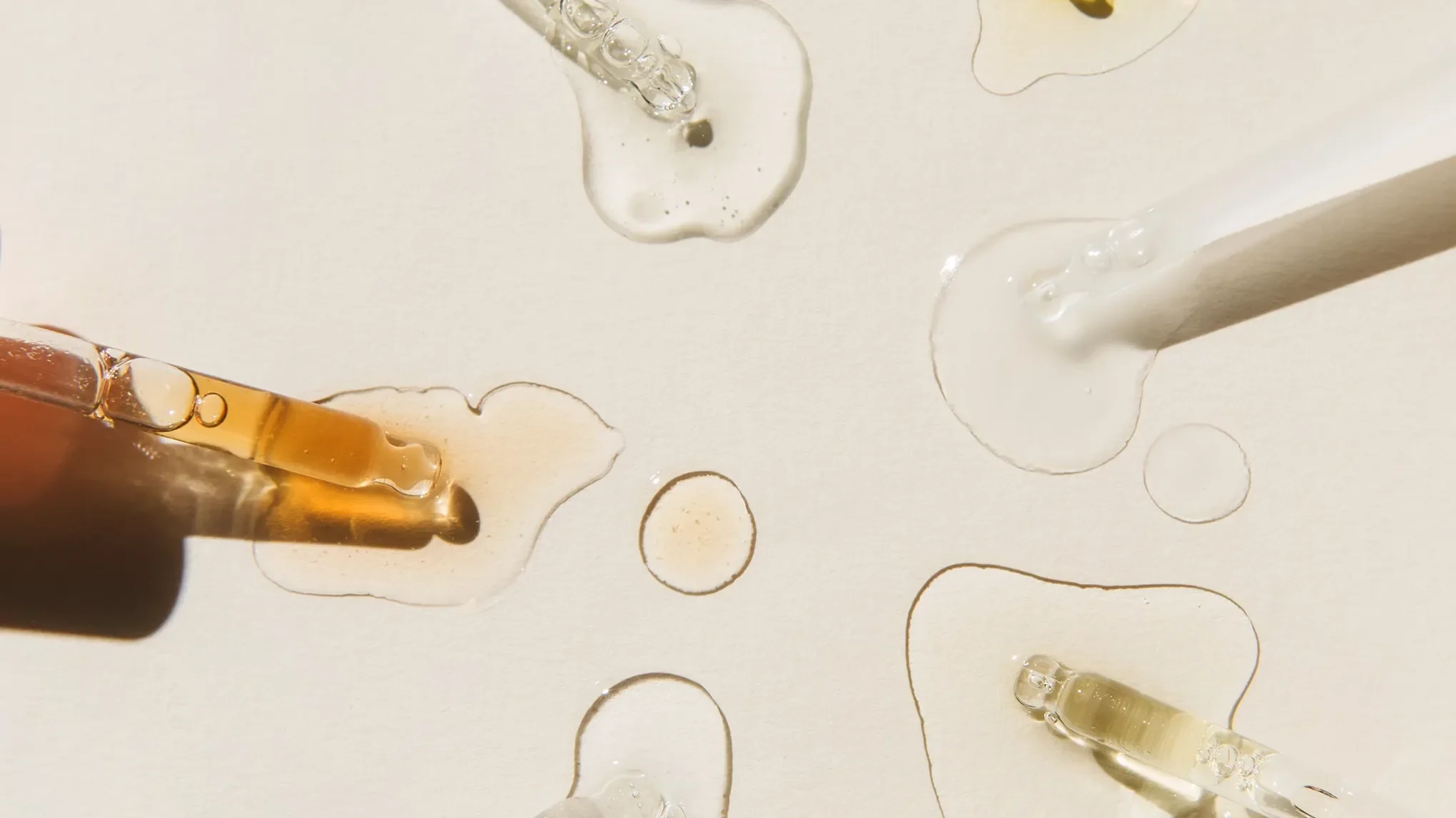 Droppar av sex olika vätskor på en beige texturerad yta i solljus