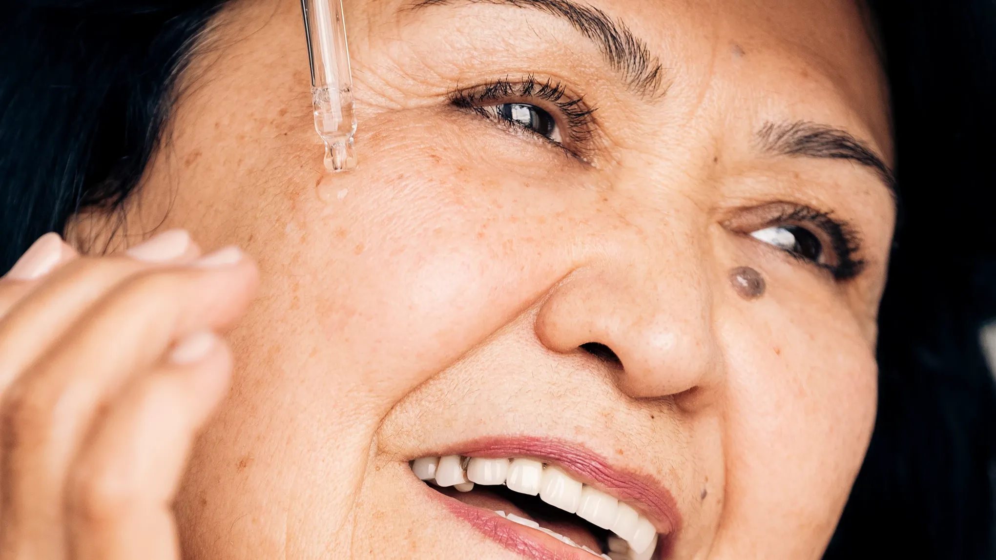 Nærbilde av en smilende eldre kvinne som påfører serum under øyet med en pipette.