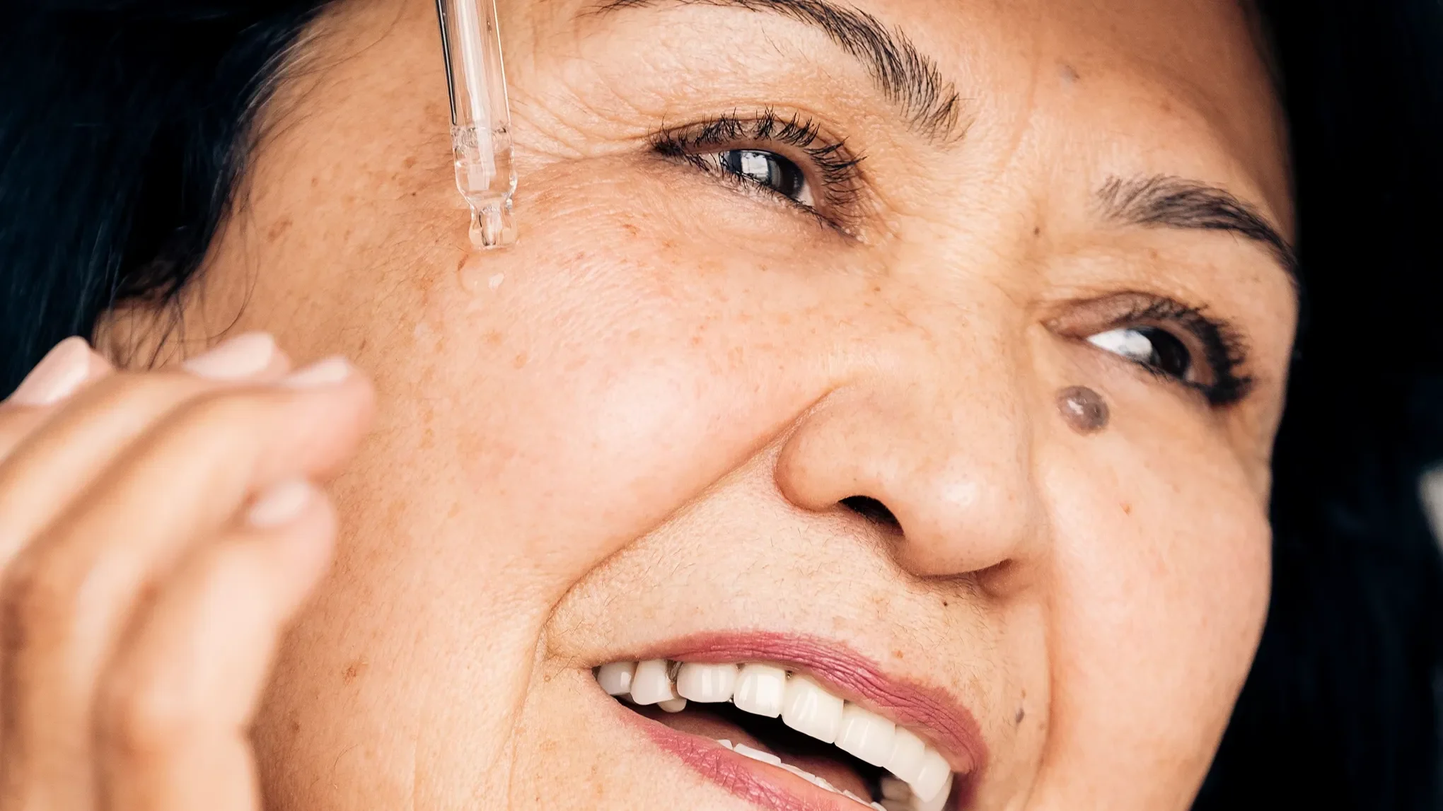 Nærbilde av en smilende eldre kvinne som påfører serum under øyet med en pipette.