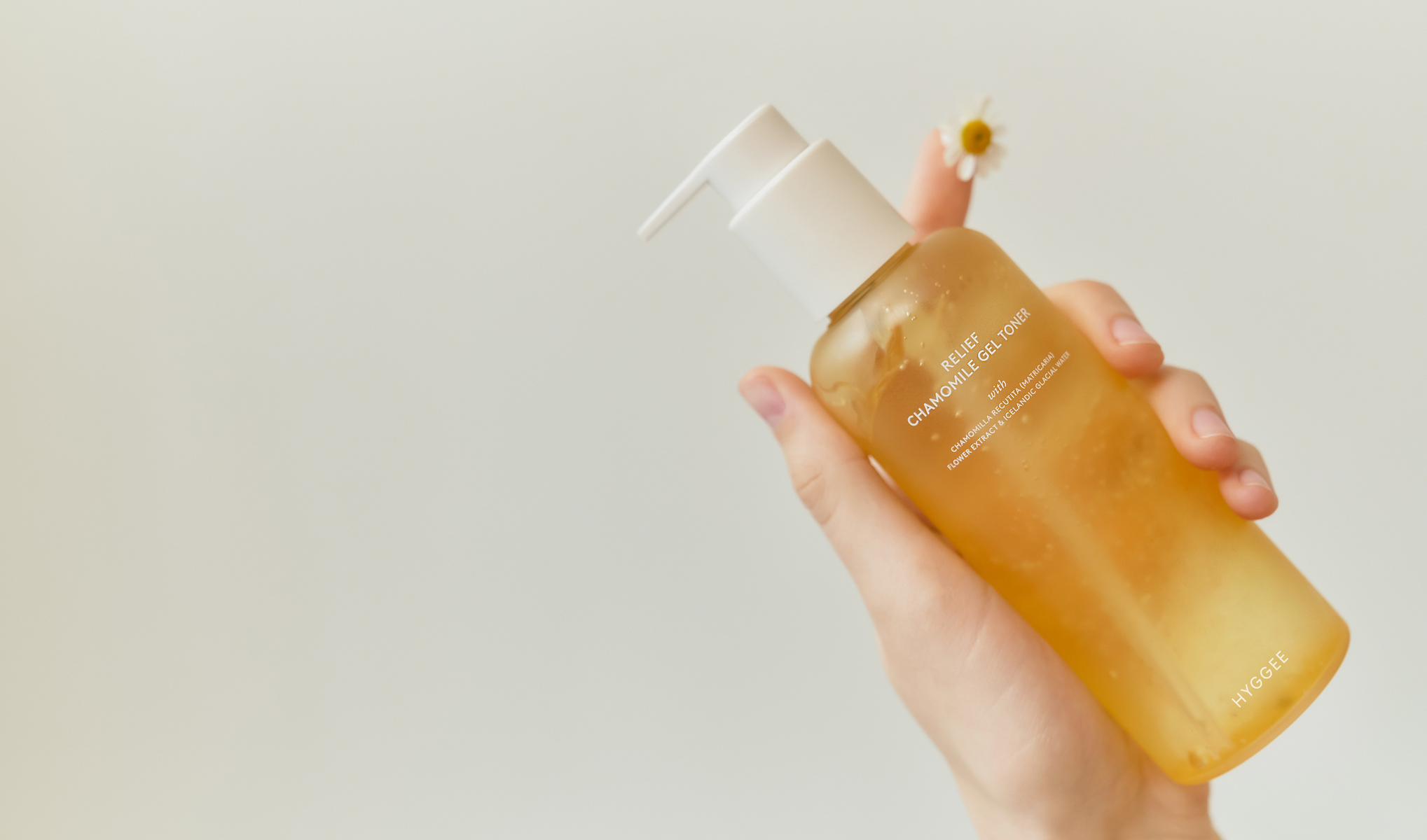 Käsi, jossa on pullo ”Relief Chamomile Gel Toner” Hyggee-merkiltä. Tausta on vaalea ja yksinkertainen, ja pullon vieressä näkyy pieni kamomillakukka.