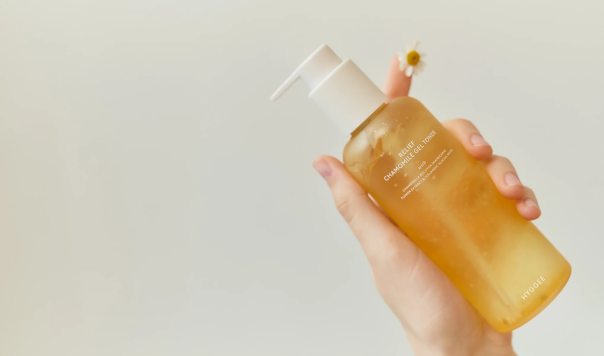 Käsi, jossa on pullo ”Relief Chamomile Gel Toner” Hyggee-merkiltä. Tausta on vaalea ja yksinkertainen, ja pullon vieressä näkyy pieni kamomillakukka.