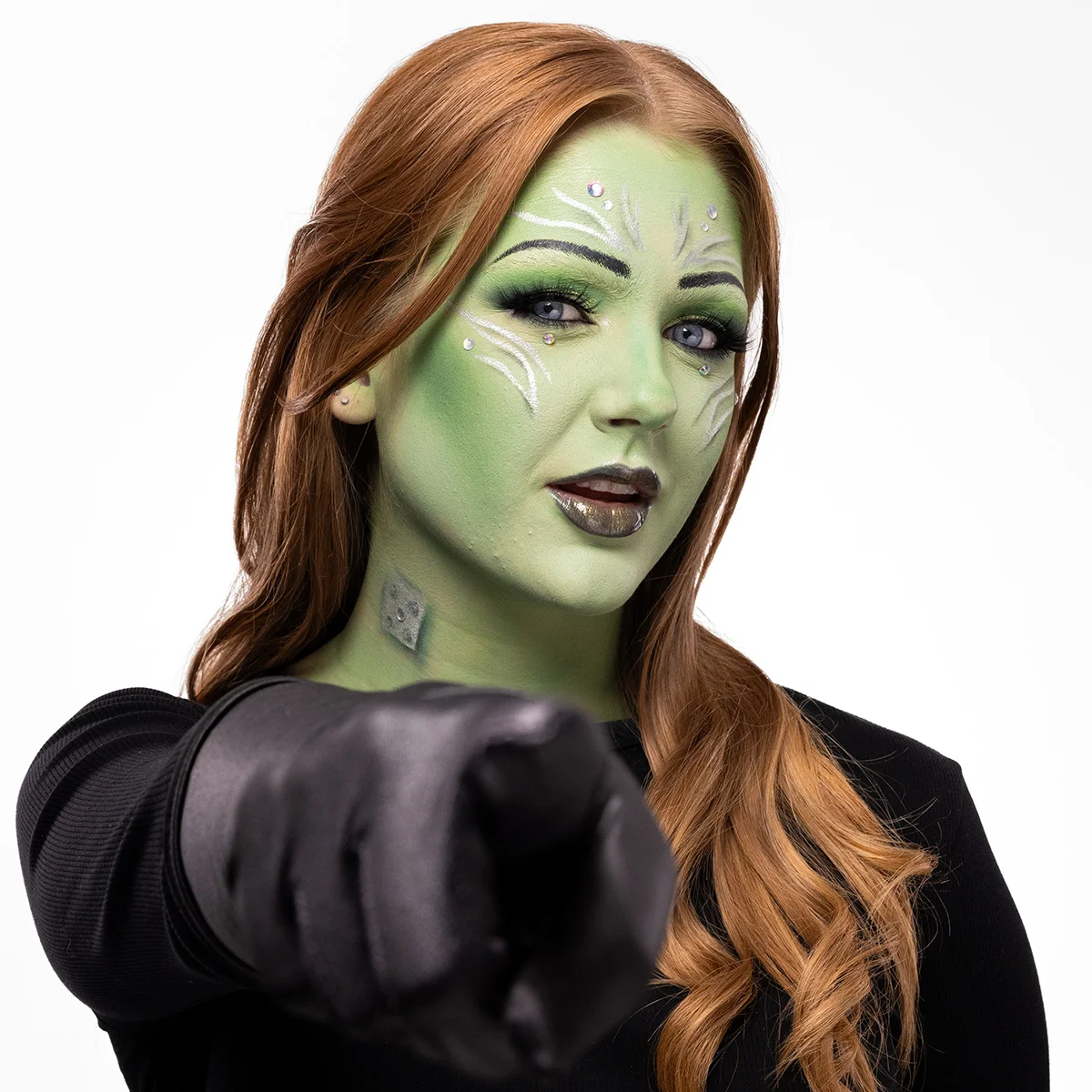 En person med grön helansiktsmakeup med silverdetaljer och små kristaller, svartglänsande läppar och svarta handskar, i en sci-fi-inspirerad look där fingret pekar mot kameran.