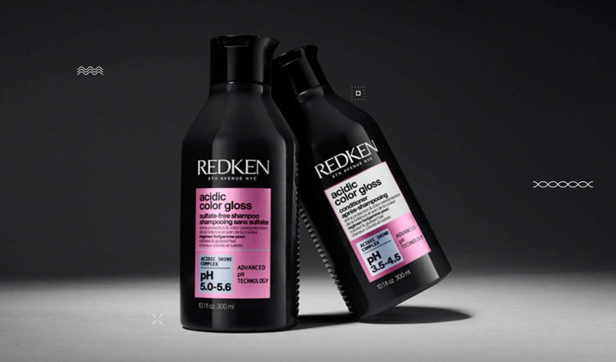 Kaksi mustaa Redken Acidic Colour Gloss -pulloa seisoo tummaa taustaa vasten. Toisessa pullossa on vaaleanpunainen etiketti ja toisessa valkoinen ja vaaleanpunainen etiketti. Pullot on sijoitettu niin, että ne ovat toisiaan kohti.
