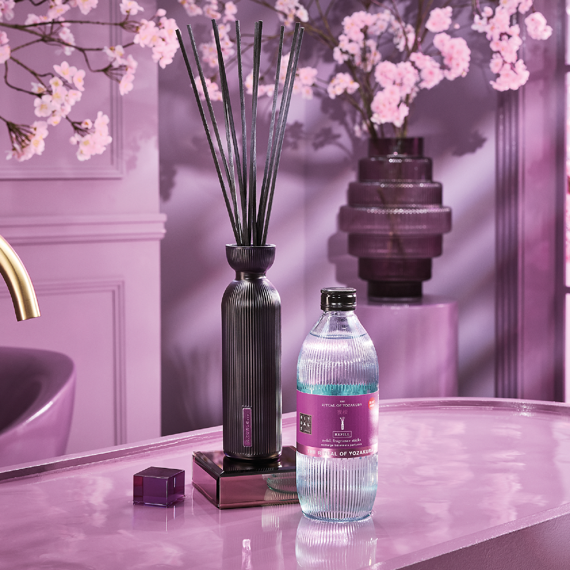Rituals-tuoksutikut mustassa pullossa ovat vaaleanpunaisella kylpyhuonepöydällä yhdessä täyttöpakkauksen kanssa, jossa lukee “THE RITUAL OF YOZAKURA”, “REFILL” ja “refill fragrance sticks”. Taustalla näkyy vaaleanpunaisia kirsikankukkia.