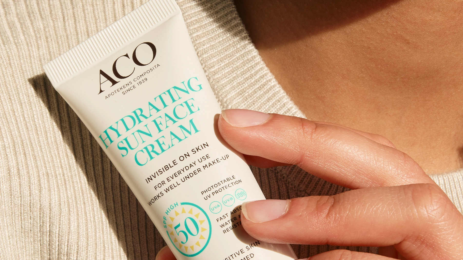 Närbild av en hand som håller en tub ACO Hydrating Sun Face Cream SPF 50 med texten “Invisible on skin”, “For everyday use works well under make-up”, “High protection 50” och “For sensitive skin unperfumed”, mot en neutral bakgrund. Fokus på solskydd för ansiktet och hudvård.