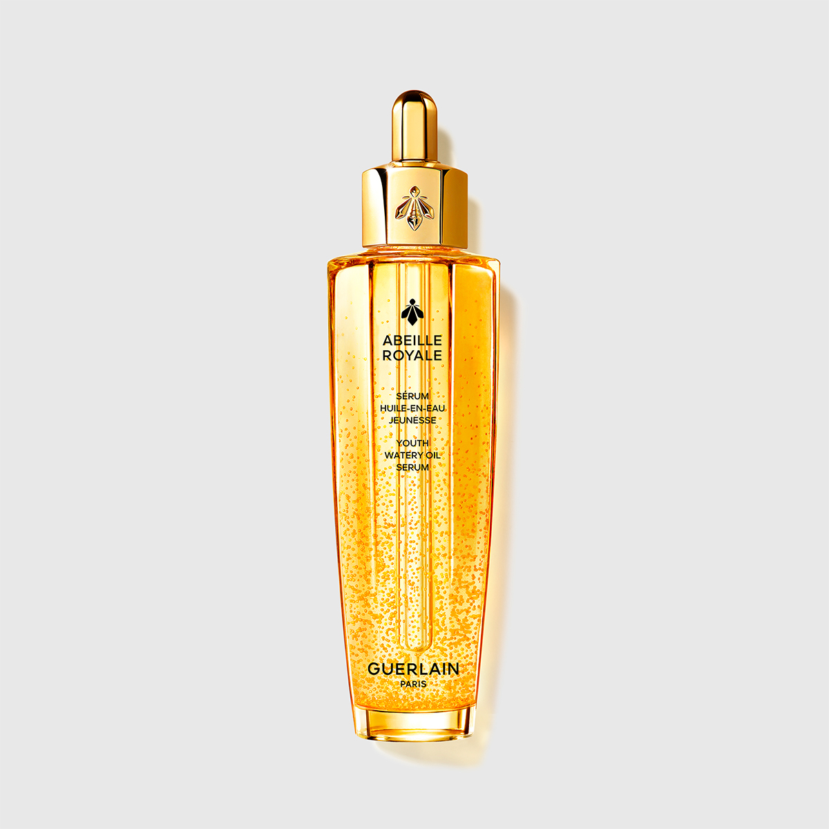 En flaske Guerlain Abeille Royale Youth Watery Oil Serum mot en hvit bakgrunn. Flasken er gyllen og gjennomsiktig, med en elegant gullhette og en pipette for påføring. Flasken har et luksuriøst og sofistikert design.