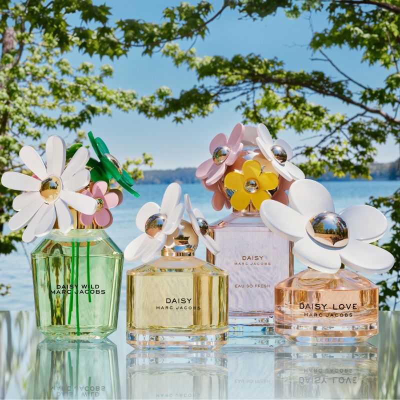 Fire parfymeflasker fra Marc Jacobs' «Daisy»-kolleksjon plassert på et utendørs bord med en innsjø og frodige trær i bakgrunnen. Flaskene har blomsterformede korker i ulike farger og er merket med «Daisy Wild», «Daisy», «Daisy Eau So Fresh» og «Daisy Love».